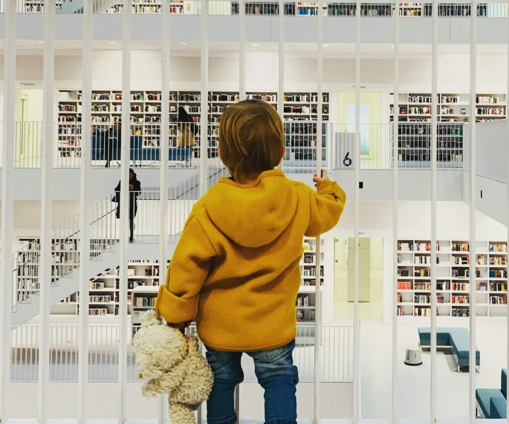 Ein Kind in einer gelben Jacke blickt mit seinem Teddybär staunend auf die weiten Regalreihen einer hellen, modernen Bibliothek. Besuch der Büchereien Wien, kuratiert für Familien auf fini.wien.